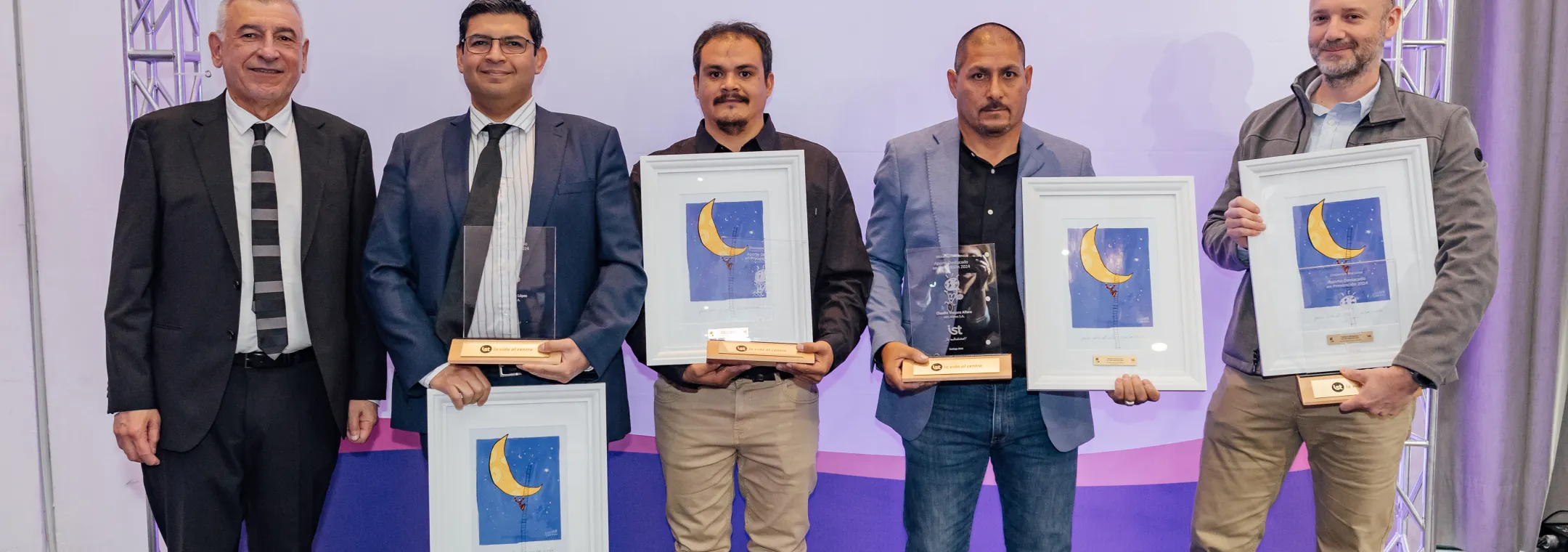 Five men pose during an award ceremony, standing in front of a backdrop with IST logo and Spanish tagline "Tu mutualidad!" Three recipients hold framed certificates featuring a crescent moon design and one also holds a trophy. The men are dressed in formal and business casual attire. The event celebrates achievements in a professional setting.