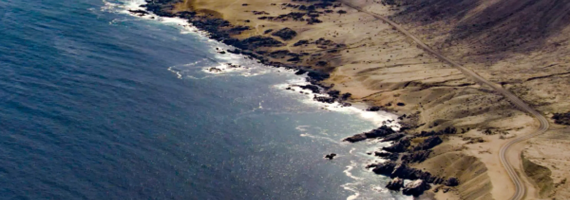 An aerial view of the coastline near Taltal, Chile. The image features a dramatic meeting of the Pacific Ocean and arid desert terrain, with rugged rocky shores and sandy hills extending into the distance. A winding dirt road follows the edge of the coast, with waves gently lapping against the shore under a hazy sky.