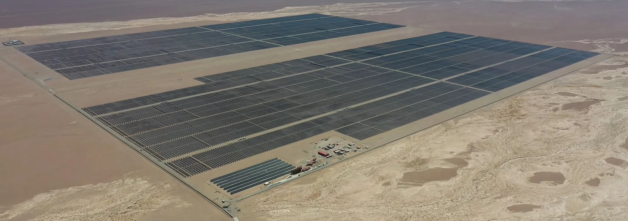 Aerial view of a large solar farm in the Atacama Desert, Chile, featuring expansive rows of dark solar panels covering vast sections of the arid landscape. The barren desert terrain surrounds the facility, with a small operations area visible near the bottom. The distant mountains and a clear blue sky can be seen in the background.