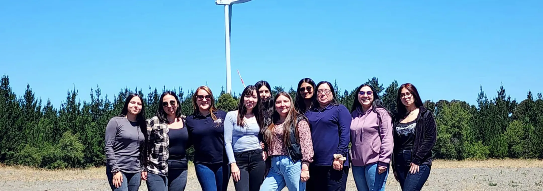 A group of women stand together in a field. Behind them is a tall, white wind turbine with red stripes at the top of the blades. The women are dressed in casual clothing such as jeans, sweaters and sneakers. They appear to be of diverse ages and ethnicities. The sky is clear and blue, and there's a line of evergreen trees in the distance. The ground is a mix of dirt and gravel. The photo conveys a sense of camaraderie in an outdoor setting.