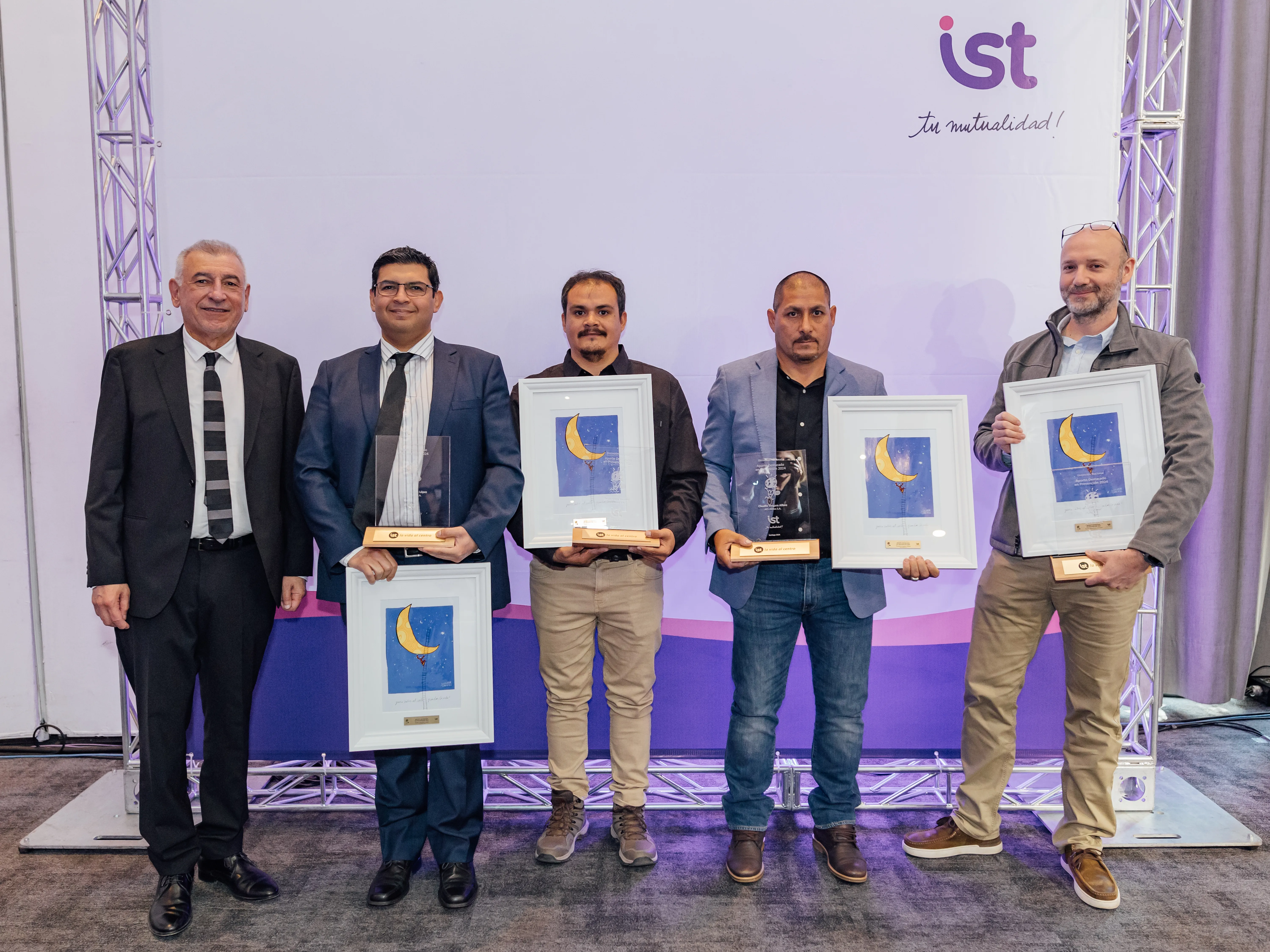 Five men pose during an award ceremony, standing in front of a backdrop with IST logo and Spanish tagline "Tu mutualidad!" Three recipients hold framed certificates featuring a crescent moon design and one also holds a trophy. The men are dressed in formal and business casual attire. The event celebrates achievements in a professional setting.