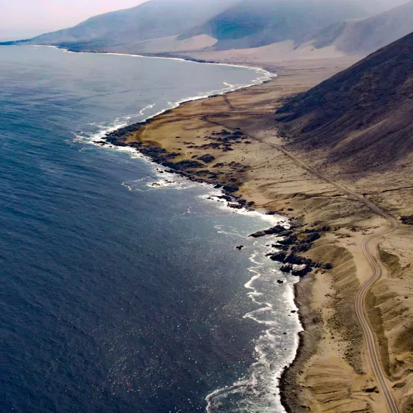 An aerial view of the coastline near Taltal, Chile. The image features a dramatic meeting of the Pacific Ocean and arid desert terrain, with rugged rocky shores and sandy hills extending into the distance. A winding dirt road follows the edge of the coast, with waves gently lapping against the shore under a hazy sky.