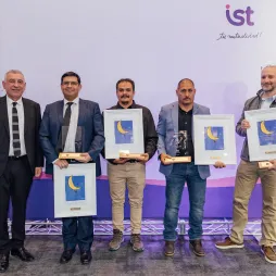 Five men pose during an award ceremony, standing in front of a backdrop with IST logo and Spanish tagline "Tu mutualidad!" Three recipients hold framed certificates featuring a crescent moon design and one also holds a trophy. The men are dressed in formal and business casual attire. The event celebrates achievements in a professional setting.