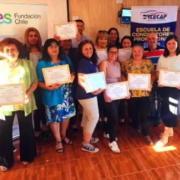 A group of women and men posing indoors during a certificate award ceremony organized by  Fundación AES Chile. Participants proudly hold certificates while standing in front of banners displaying the AES logo and the text "Escuela de Conductores Profesionales" (Professional Drivers' School). The room has wooden walls, a window allowing natural light, and polished wooden flooring.