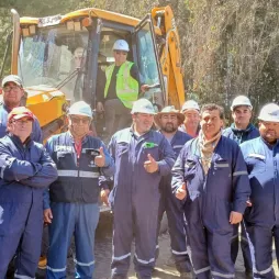 A group of construction workers wearing blue uniforms, hard hats, and safety gear, standing outdoors in front of a yellow bulldozer. One worker is seated inside the bulldozer's cabin. The background includes trees and greenery under a bright sky. The workers are smiling, some giving thumbs-up, creating a positive and collaborative atmosphere.