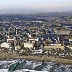 Aerial view of the Ventanas power plant located on the Chilean coast, featuring four industrial units with chimneys emitting smoke. The plant is situated near the shoreline with ocean waves in the foreground. Surrounding the facility is a town with houses, roads, and greenery, alongside industrial infrastructure and a river cutting through the landscape.