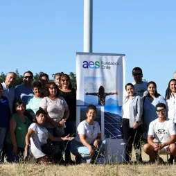 A group of people poses in front of a wind turbine at a renewable energy site during a community event organized by AES Fundación Chile. A banner with the AES logo and images of solar panels and a person with open arms is displayed in the center. The setting is an open field with clear blue skies, and additional wind turbines are visible in the background