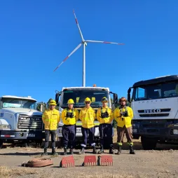 Five firefighters wearing yellow protective gear, helmets, and safety goggles stand in front of three large trucks, including a tanker and a heavy-duty vehicle, with firefighting tools placed on the ground. A wind turbine towers in the background against a clear blue sky, indicating a renewable energy site. The team stands confidently, ready for emergency response.