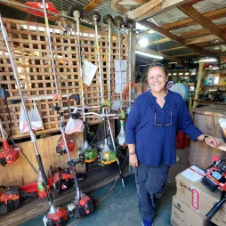  A woman stands smiling in a display of various gardening tools and equipment. The space features a wooden interior with neatly arranged trimmers, blowers and other tools on shelves. The atmosphere reflects a local business specializing in gardening and landscaping supplies.