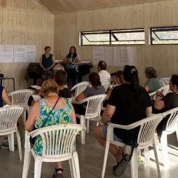 A group of people seated on white plastic chairs inside a wood-paneled room attend a presentation. Two women stand at the front near a projector screen displaying text. An AES Fundación Chile banner is visible on the left. Attendees are engaged, some taking notes. Natural light enters through horizontal windows, and refreshments are set up on a table in the background