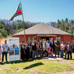 A group of people stands in front of a wooden rural house with a thatched roof, posing for a photo. A Mapuche flag is raised on a pole nearby. The group includes men, women, and children, some wearing traditional clothing. An AES Fundación Chile banner is displayed, featuring images of solar panels and a person with open arms. The background consists of green hills and trees under a clear blue sky.