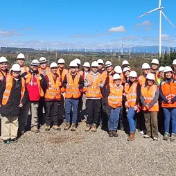 A large group of people wearing orange safety vests and white hard hats stand together at a wind farm site, posing for a photo. The background features multiple wind turbines on rolling hills under a bright blue sky. The group is diverse, with individuals of different ages and genders, standing on a gravel-covered ground with trees on one side.