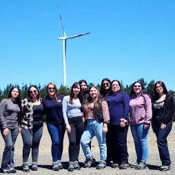 A group of women stand together in a field. Behind them is a tall, white wind turbine with red stripes at the top of the blades. The women are dressed in casual clothing such as jeans, sweaters and sneakers. They appear to be of diverse ages and ethnicities. The sky is clear and blue, and there's a line of evergreen trees in the distance. The ground is a mix of dirt and gravel. The photo conveys a sense of camaraderie in an outdoor setting.